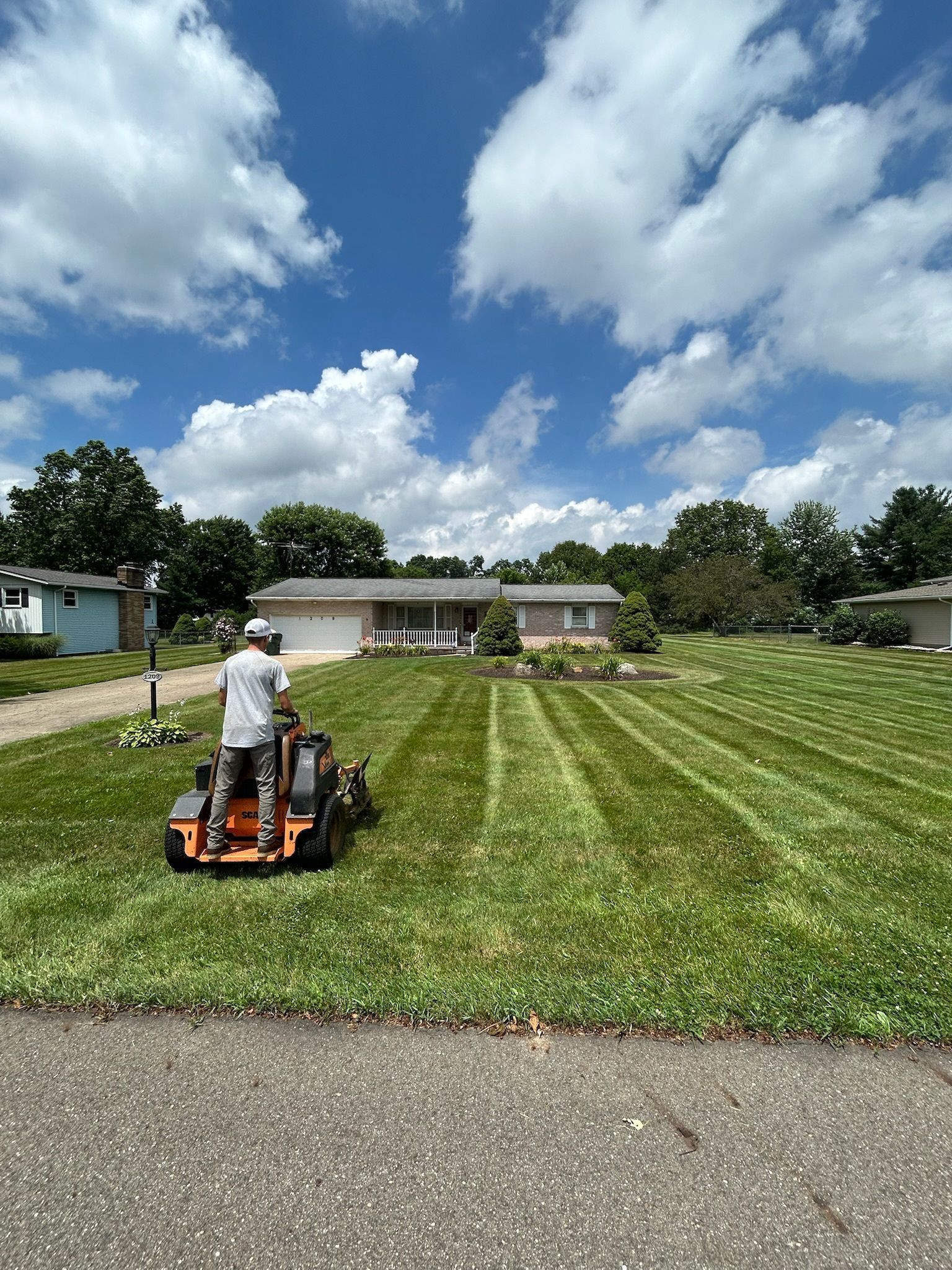 Person mowing a lawn with a riding mower, creating stripes. Sunny day with a house and trees in the background.