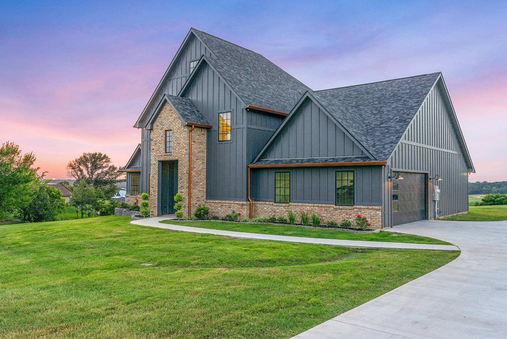 A large house with a gray siding and a garage is sitting on top of a lush green field.