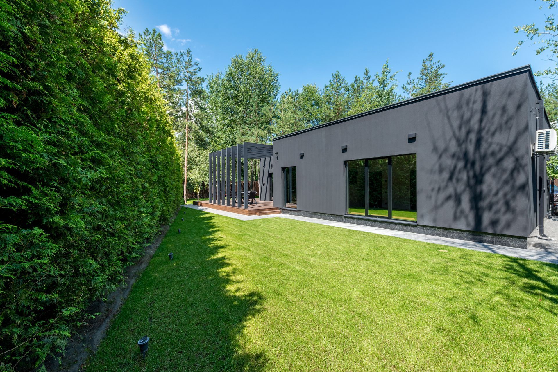 Modern gray house with large windows beside a bright green lawn and tall hedges under a blue sky