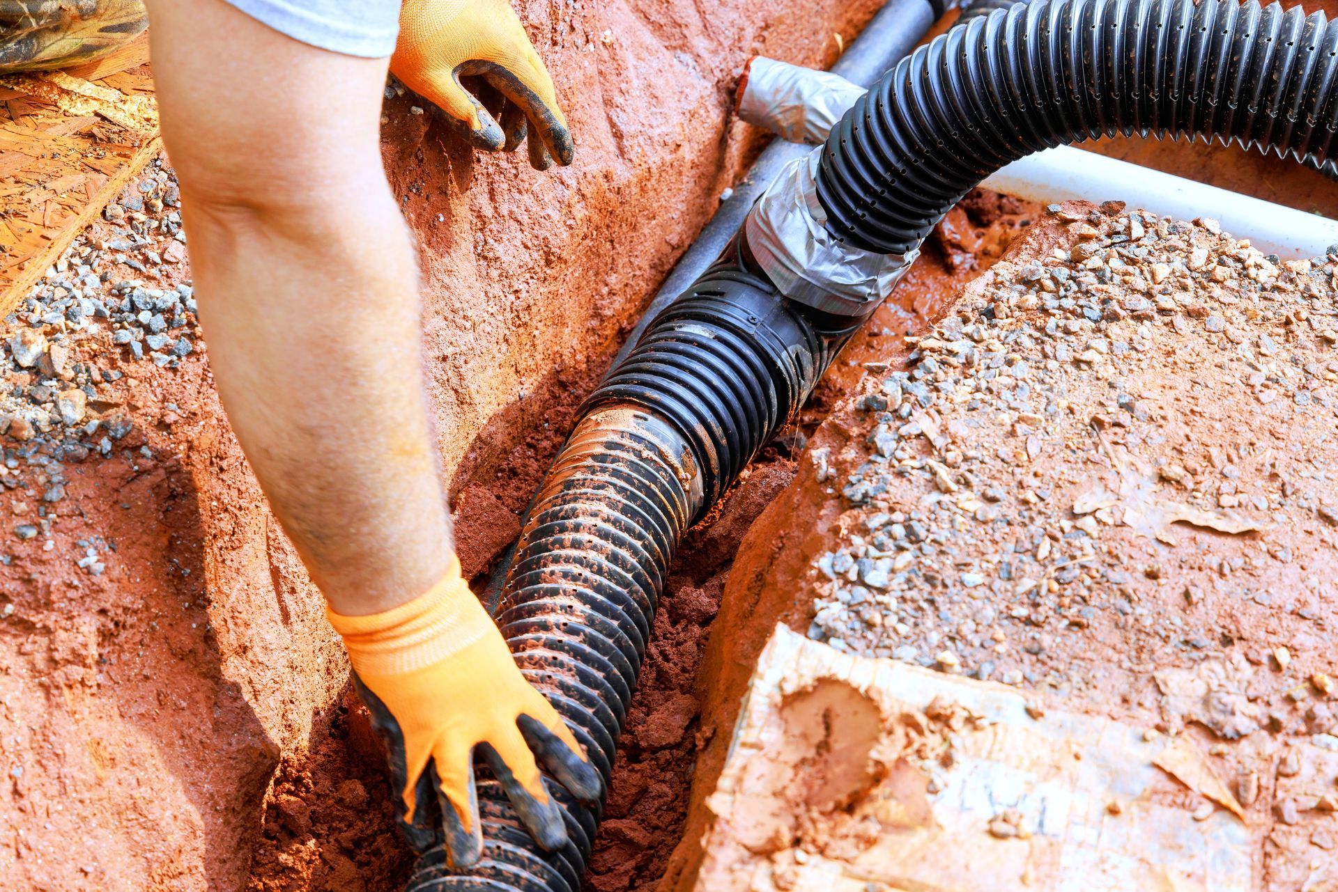 Worker guiding a black corrugated pipe into a trench with orange-gloved hands