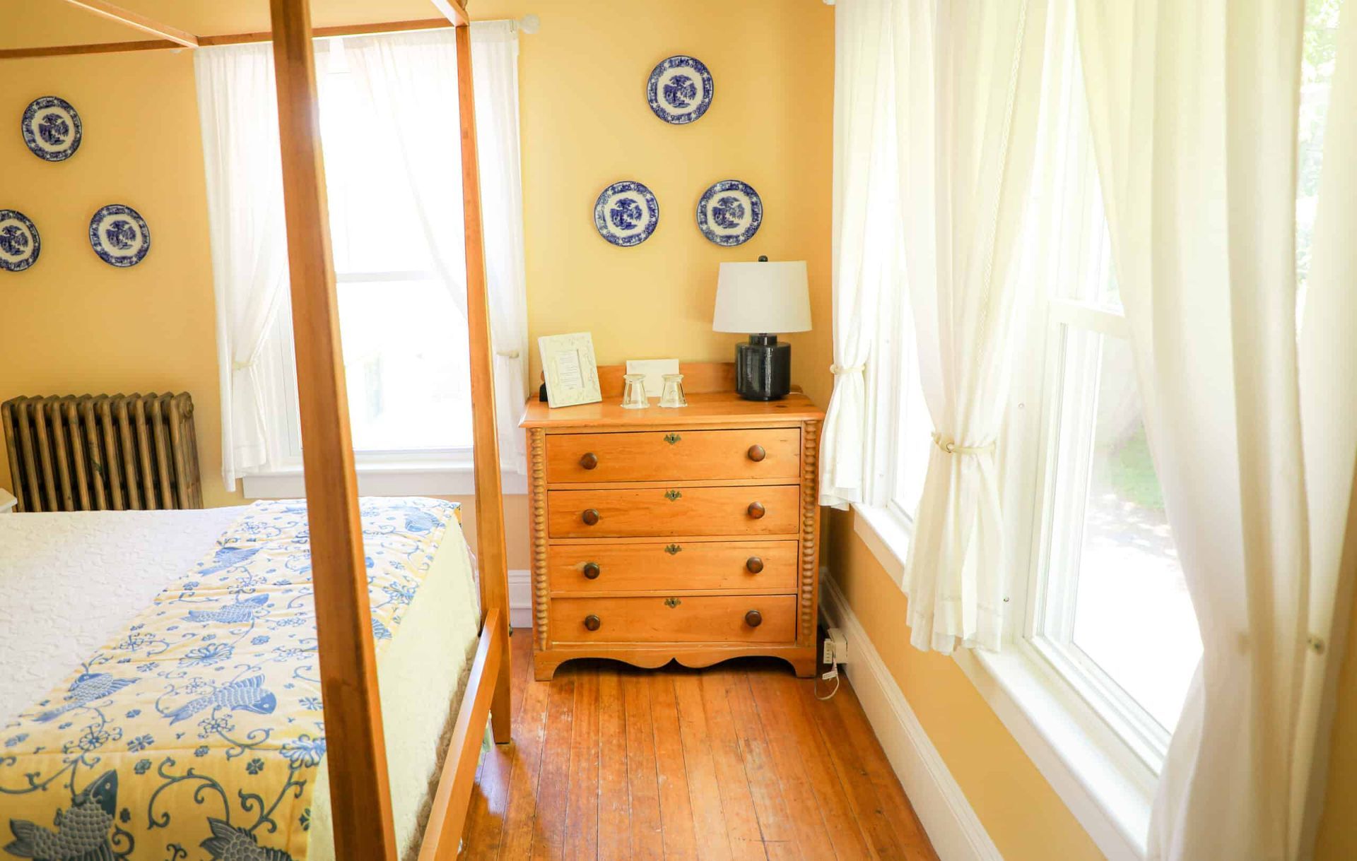 A bedroom with a four poster bed , dresser , lamp and window.