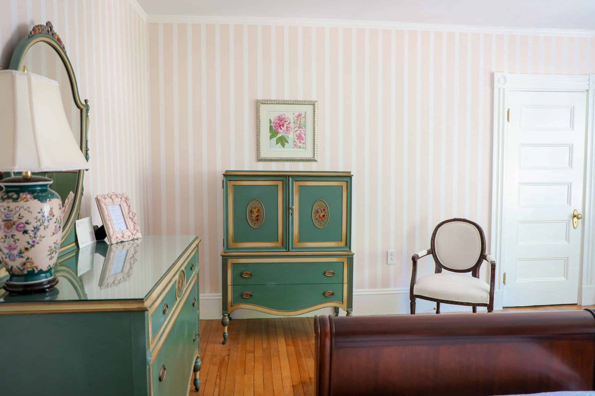 A bedroom with a dresser , chair , lamp and striped wallpaper.