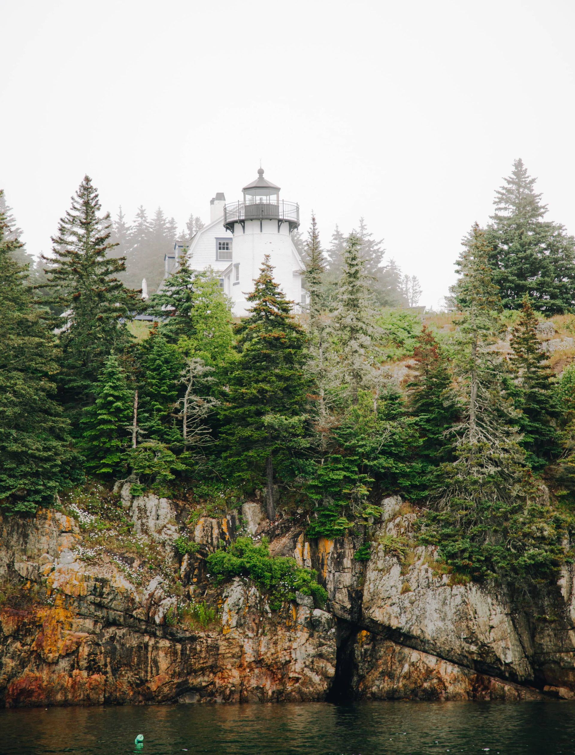 A lighthouse is sitting on top of a rocky cliff next to a body of water surrounded by trees.