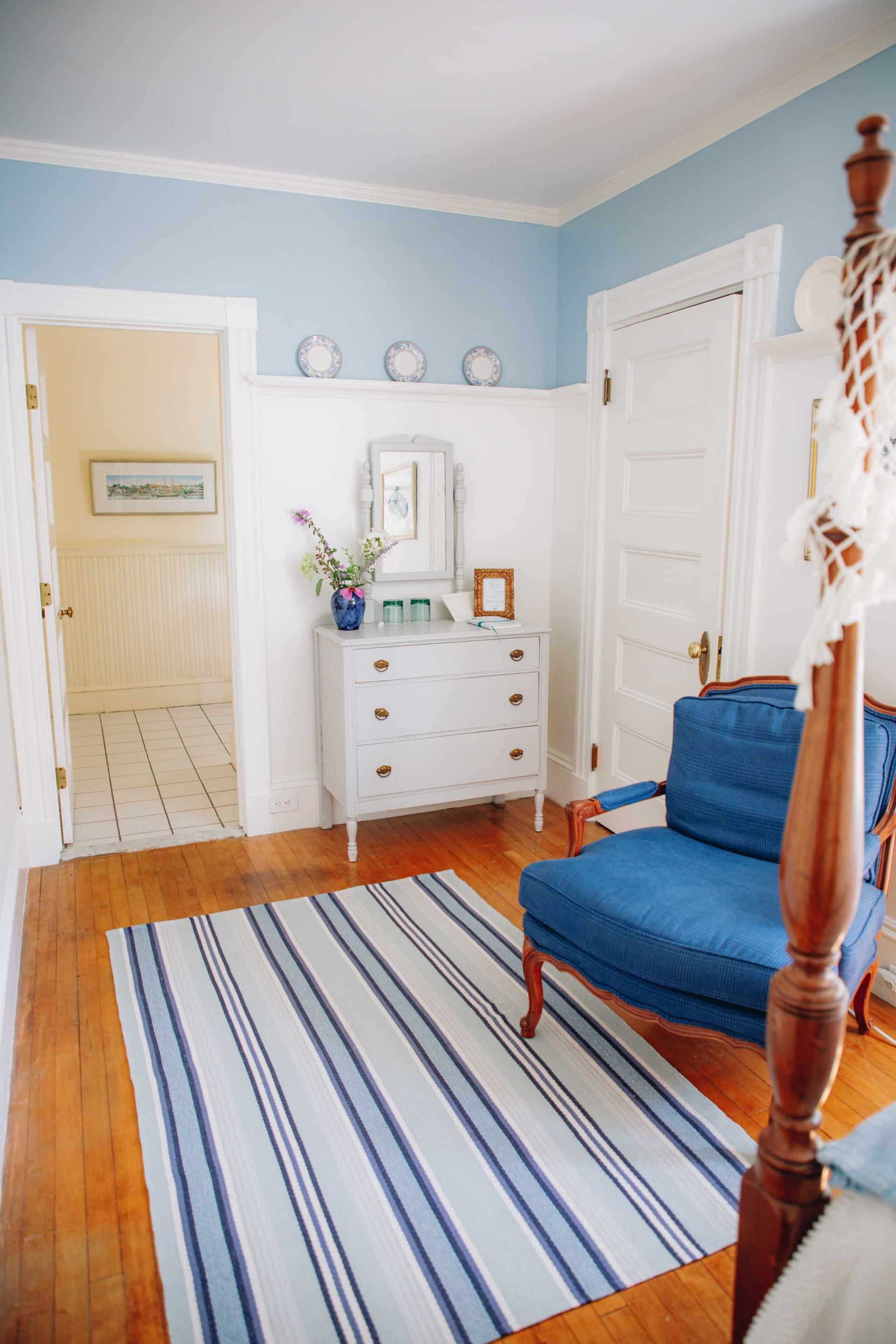 A bedroom with blue walls , a blue chair , a striped rug , a dresser and a mirror.