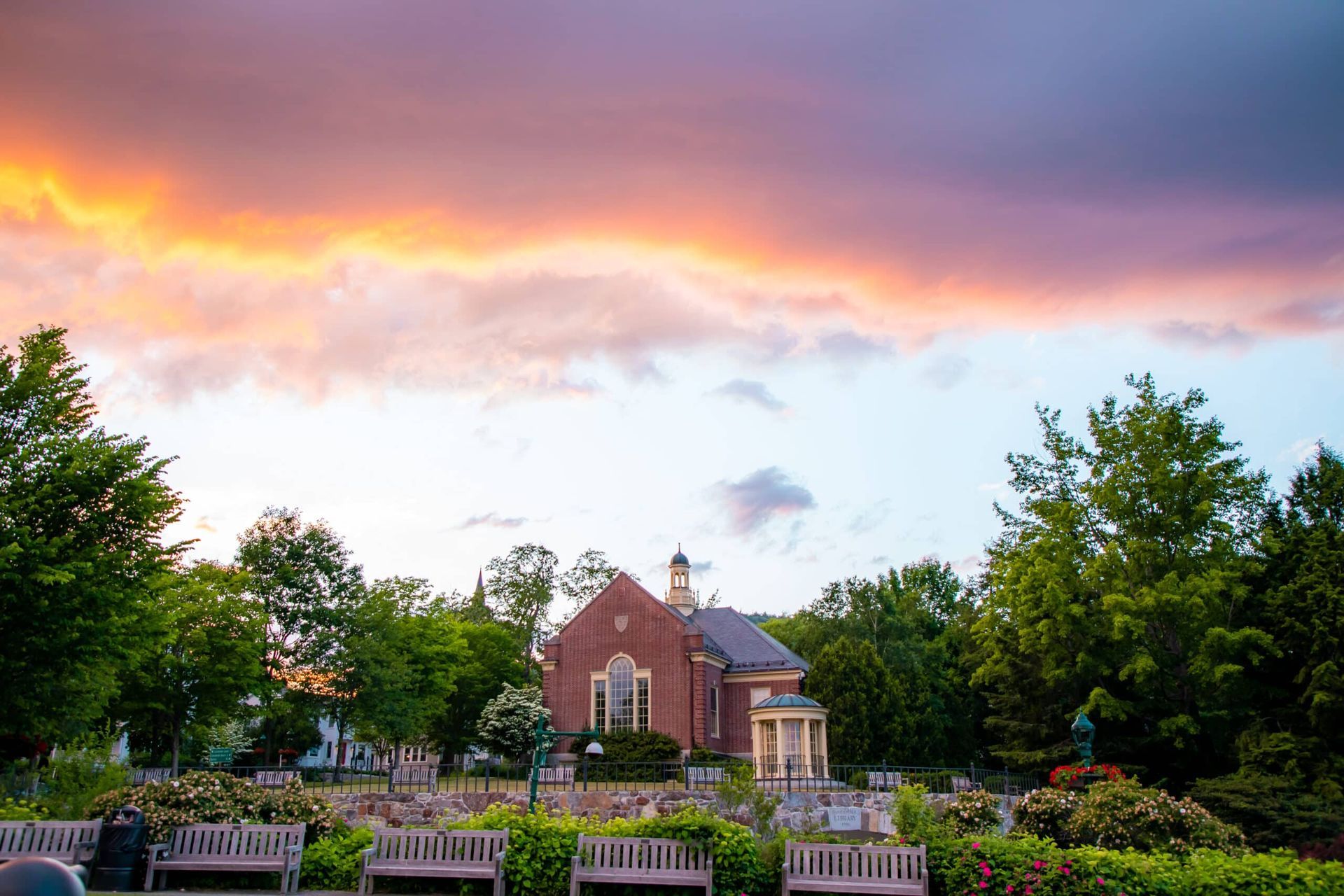 A brick building with a sunset in the background is surrounded by trees and benches.