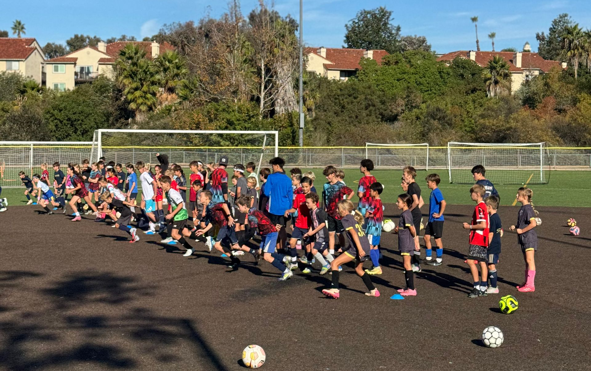 Soccer players on a field, practicing drills. Several players squatting, awaiting instruction, with goals in background.
