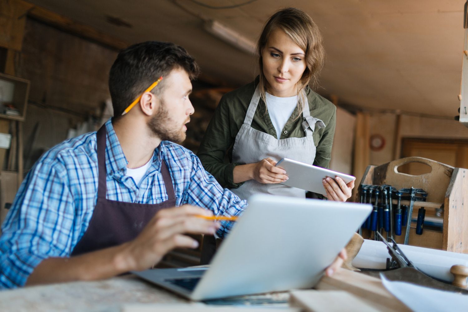 Man and woman in woodshop, working on a laptop and tablet, reviewing plans, both wearing aprons.