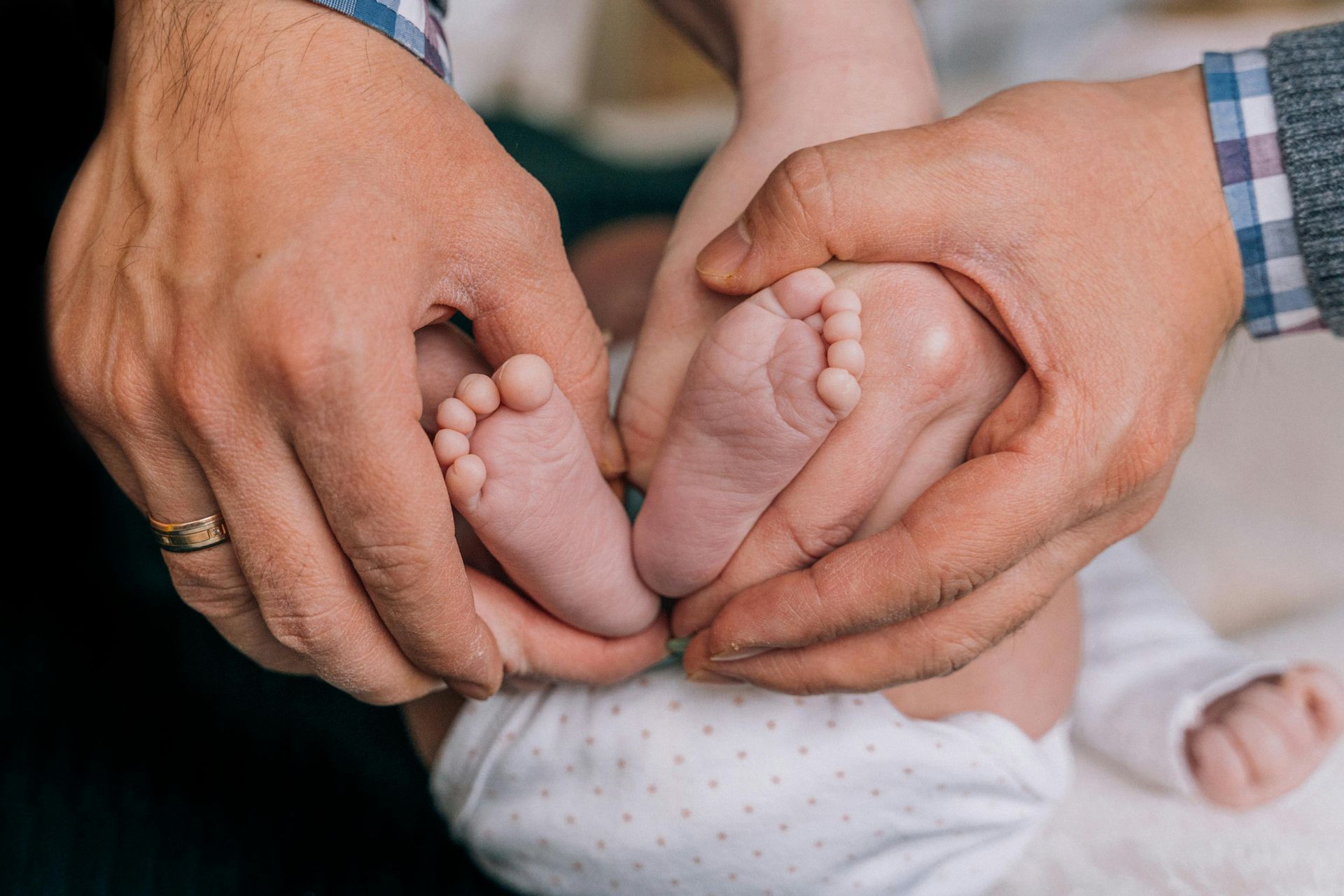 Hands holding the tiny feet of a baby, the baby is wearing a white onesie.