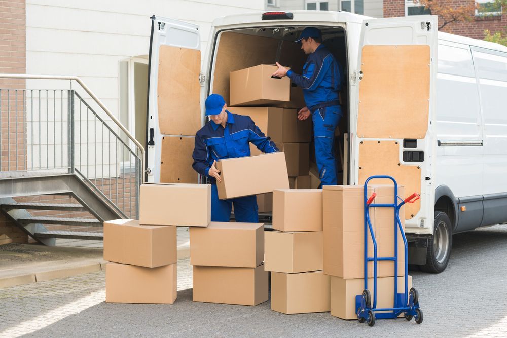 Movers in blue uniforms loading cardboard boxes into a white van outside a building.