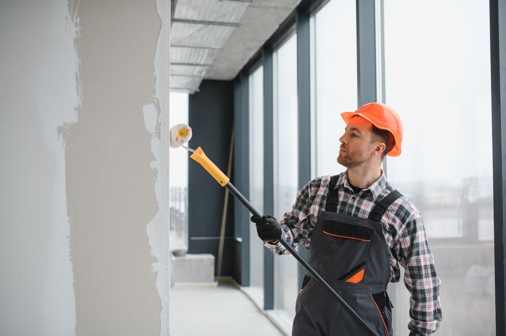 Construction worker in orange hard hat painting a wall with a roller. Interior setting, large windows.
