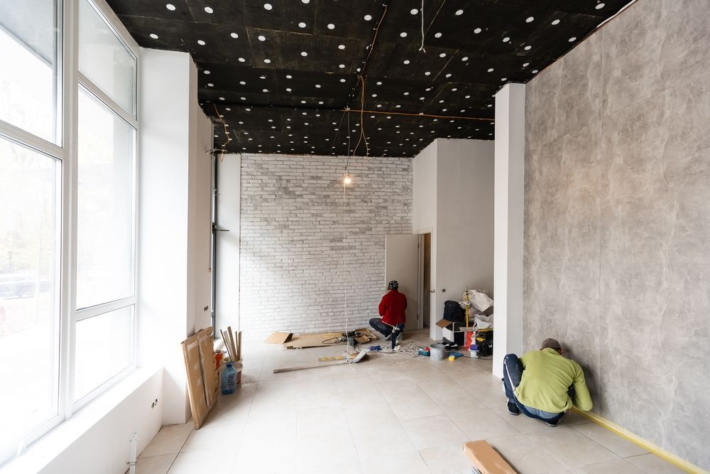 Interior renovation in progress, two workers, exposed ceiling, unfinished walls, large windows.