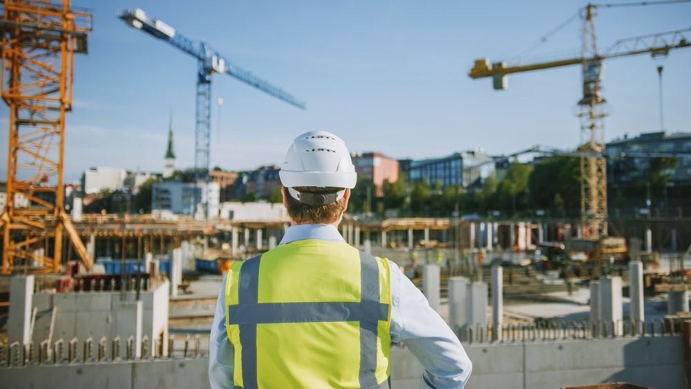Construction worker in white hard hat and yellow vest surveys a construction site with cranes and buildings.