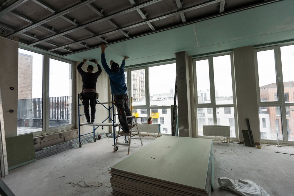 Two workers installing drywall ceiling in a room with large windows.