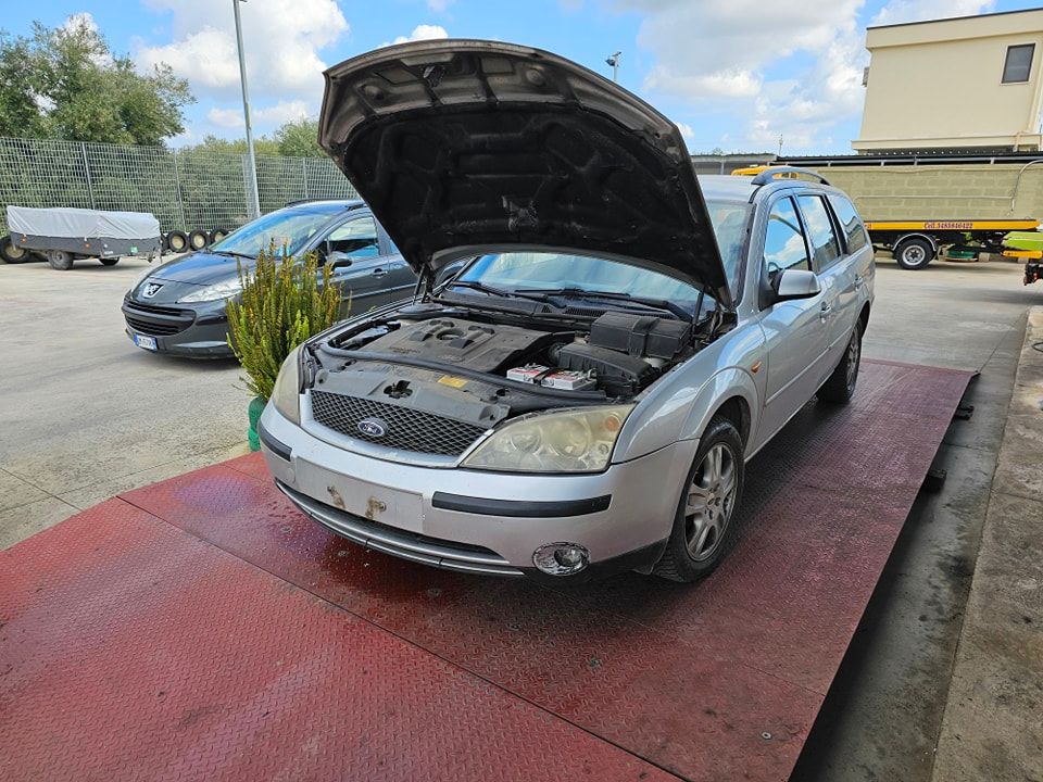 Una Ford Mondeo station wagon color argento, con il cofano aperto, parcheggiata su una piattaforma metallica arrugginita in un parcheggio all'aperto.