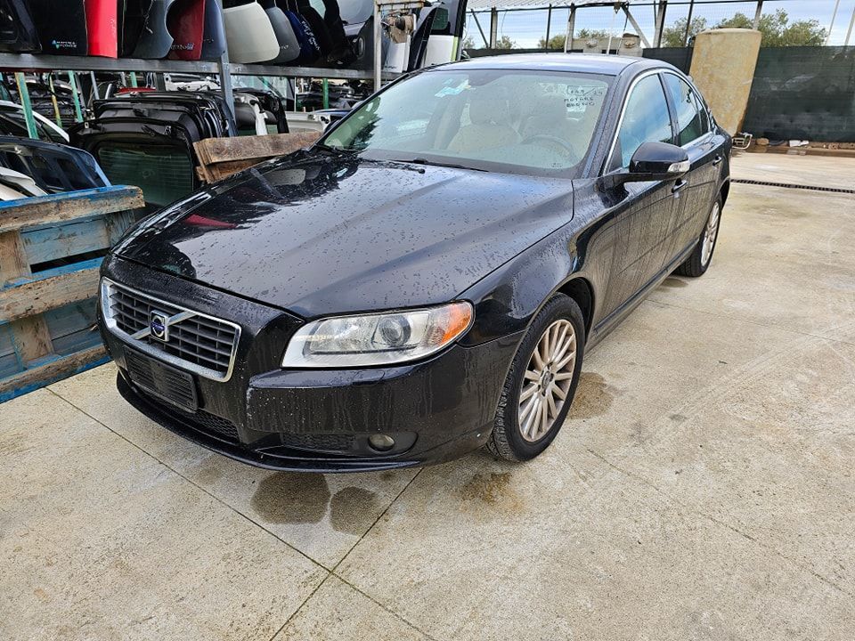 Una berlina Volvo nera parcheggiata all'aperto su una superficie di cemento, con ricambi auto su degli scaffali sullo sfondo.