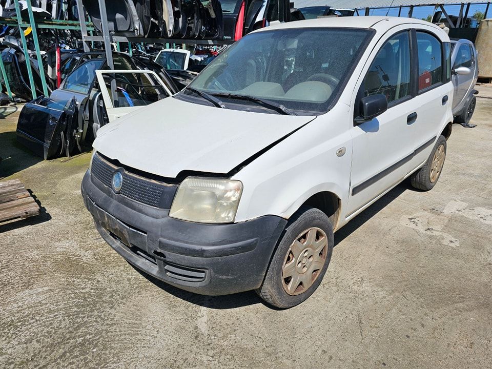 Una Fiat Panda bianca parcheggiata in una discarica a cielo aperto piena di vari pezzi di ricambio e telai di veicoli.