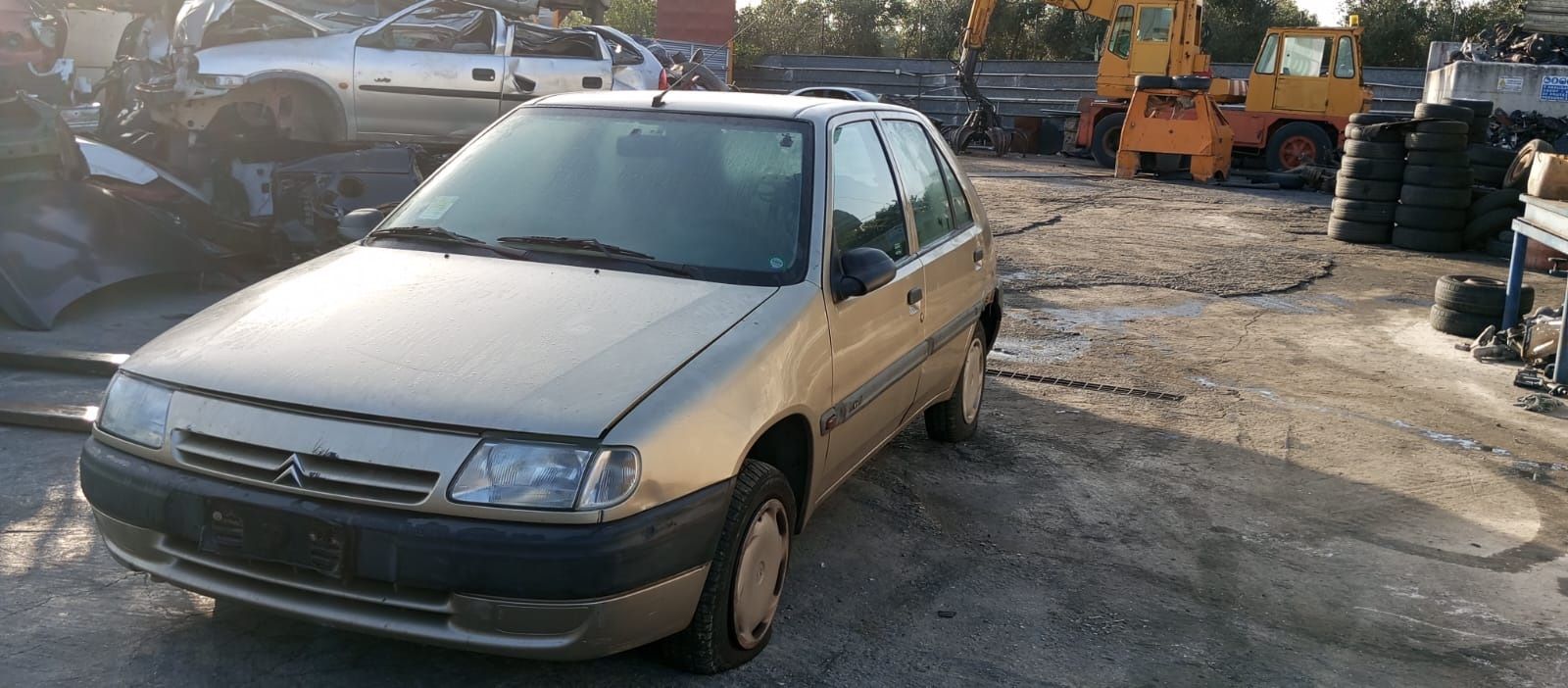 Una Citroën hatchback a quattro porte color oro parcheggiata in un deposito di rottami, con delle attrezzature sullo sfondo.
