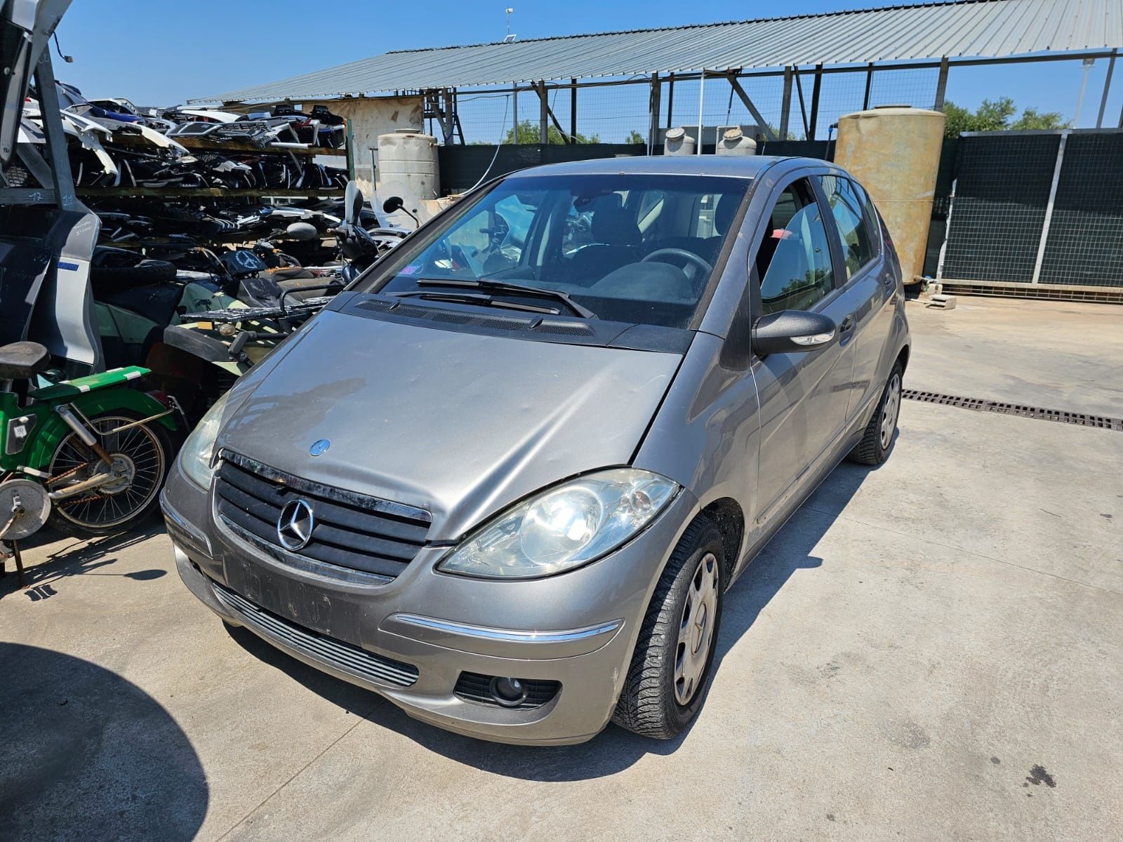 Una Mercedes-Benz Classe A hatchback color argento parcheggiata in un deposito di autodemolizioni in una giornata di sole.