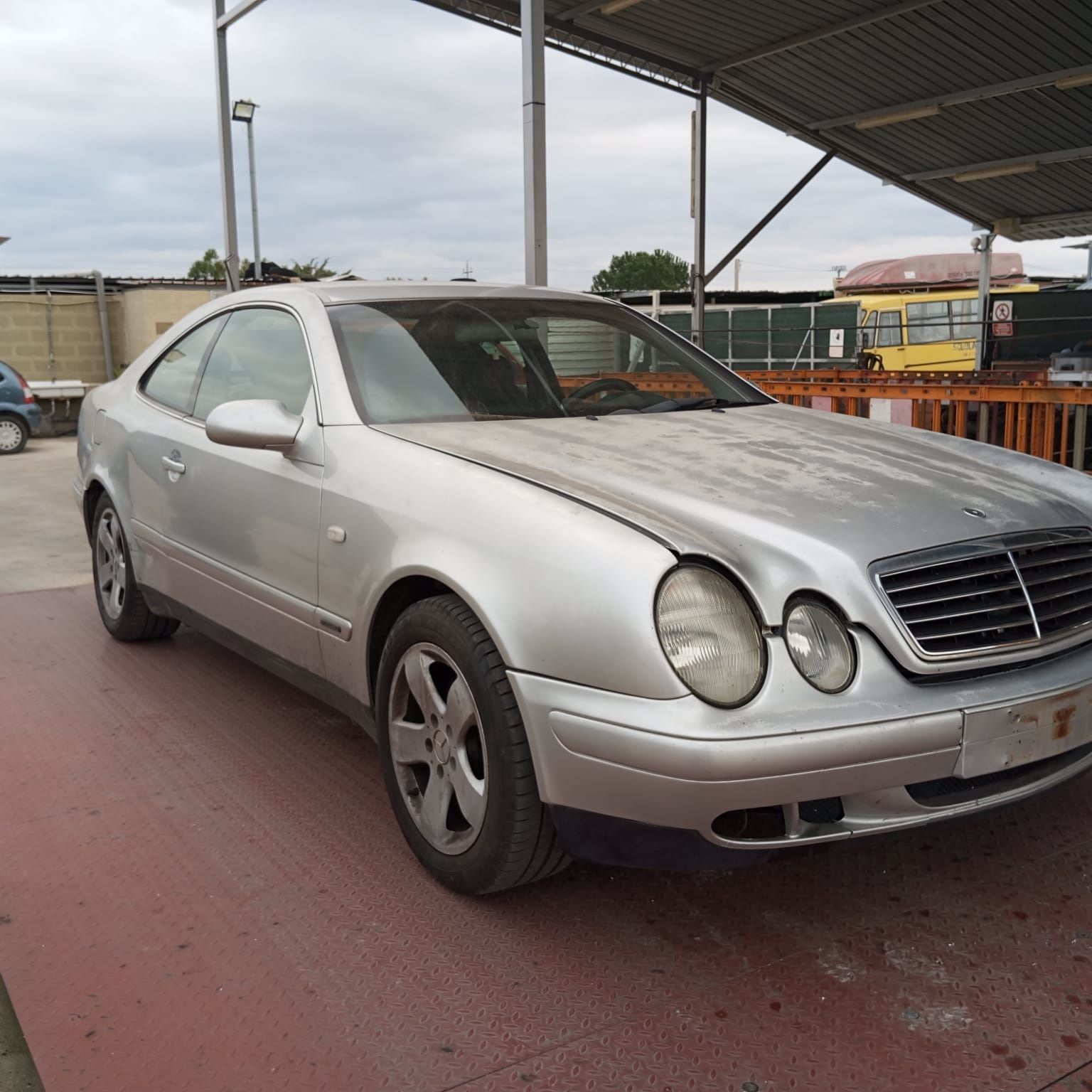 Una Mercedes-Benz CLK coupé color argento parcheggiata su una superficie rossastra sotto una tettoia metallica.