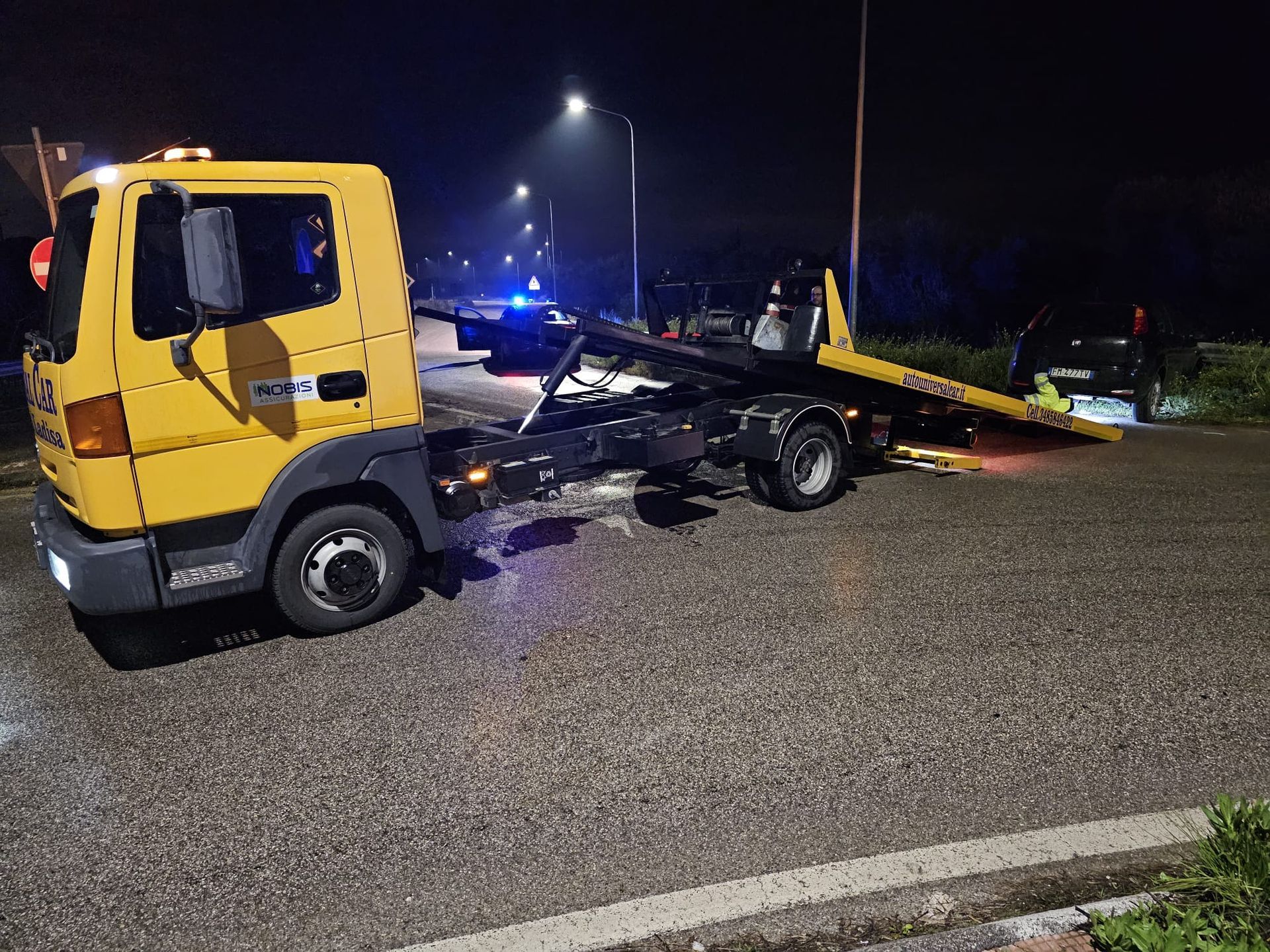 Un carro attrezzi giallo con pianale ribassato, parcheggiato di notte su una strada buia e bagnata, con la rampa abbassata verso un veicolo scuro.
