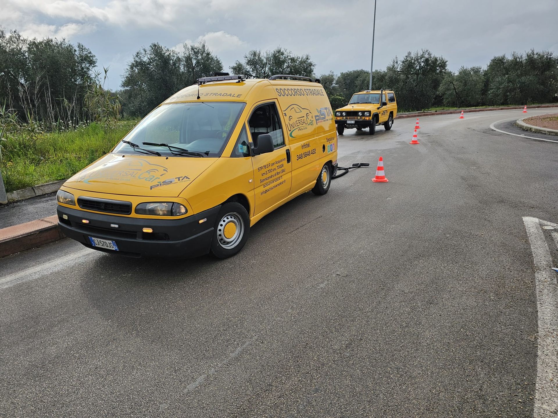 Un furgone giallo parcheggiato su una strada asfaltata con coni stradali e un altro veicolo di servizio giallo nelle vicinanze.