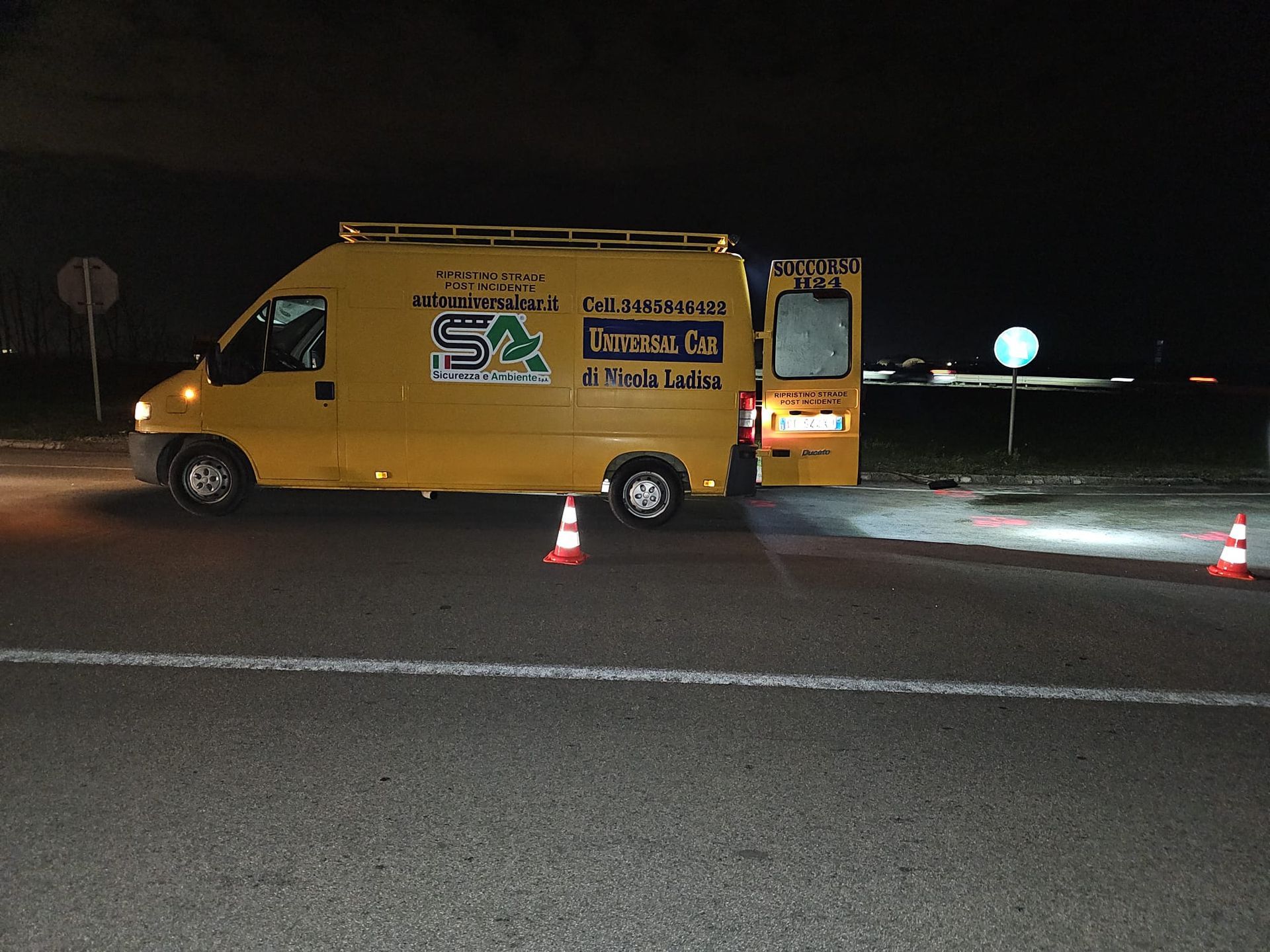 Un furgone giallo parcheggiato sul ciglio di una strada buia con il portellone posteriore aperto e due coni stradali arancioni posizionati nelle vicinanze.