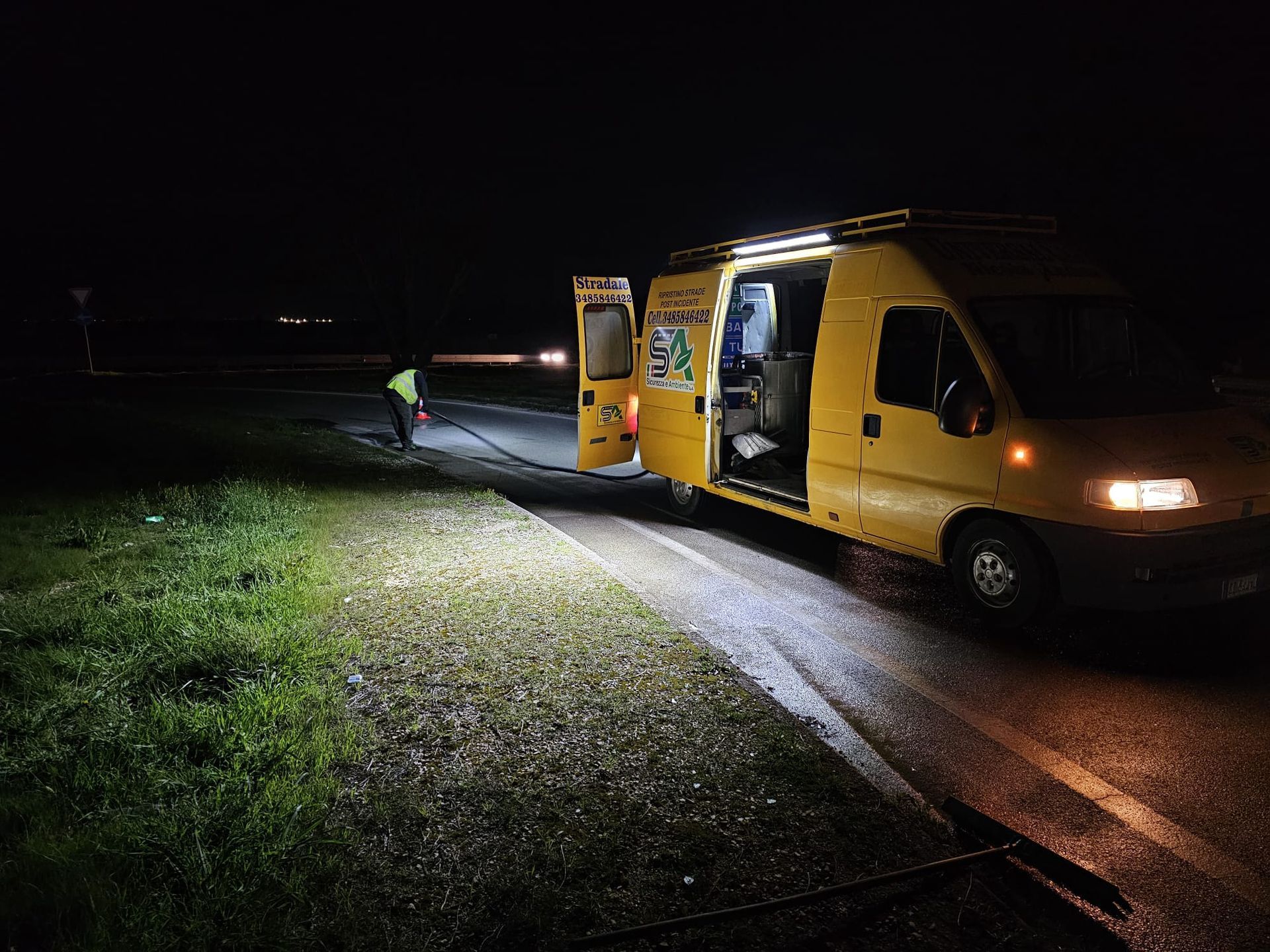 Un furgone di servizio giallo brillante parcheggiato di notte sul ciglio di una strada buia, con un tecnico al lavoro vicino allo sportello laterale aperto.