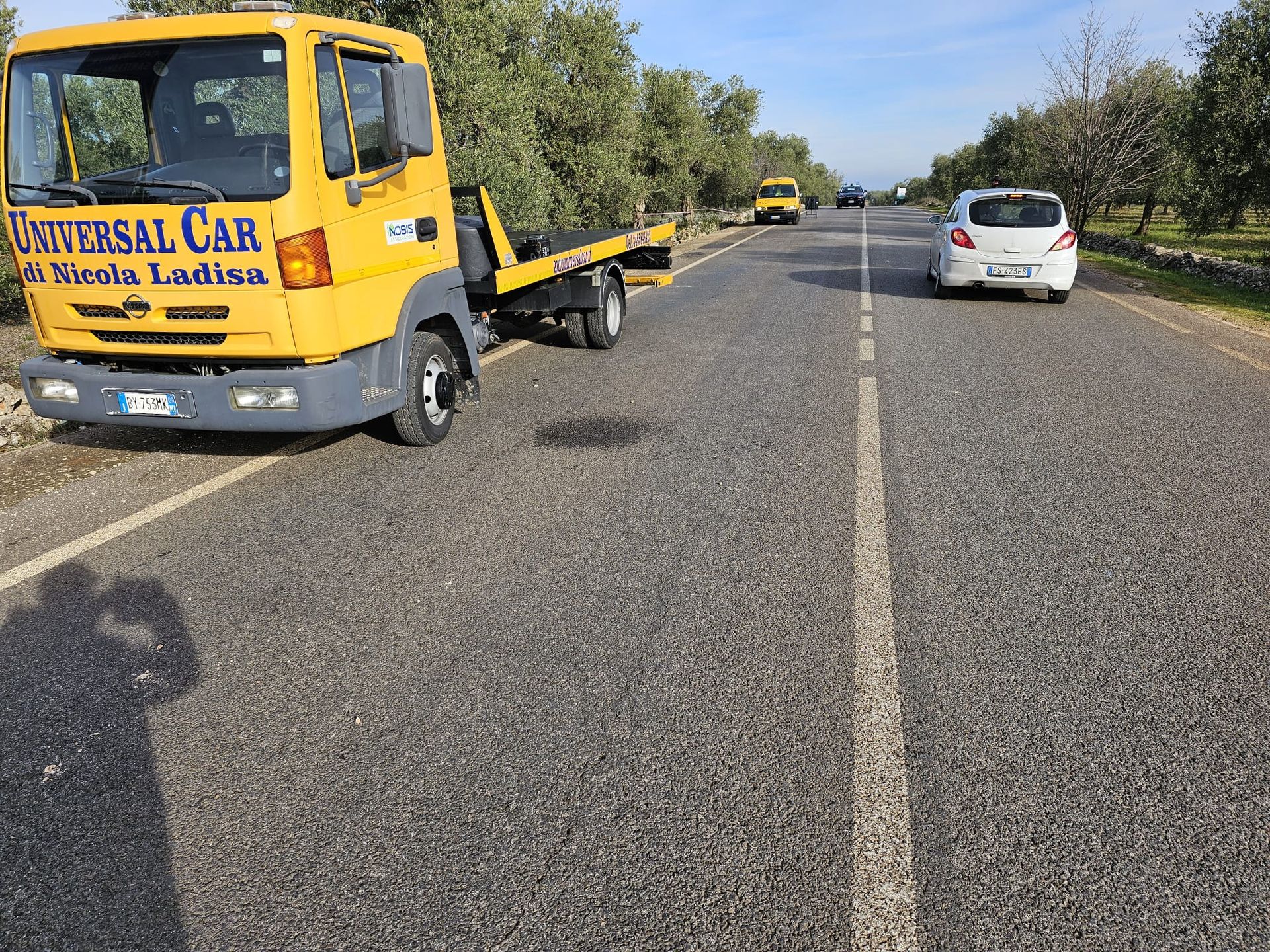 Un carro attrezzi giallo con pianale è parcheggiato sul ciglio di una strada rurale, con un'auto bianca davanti e alberi su entrambi i lati.