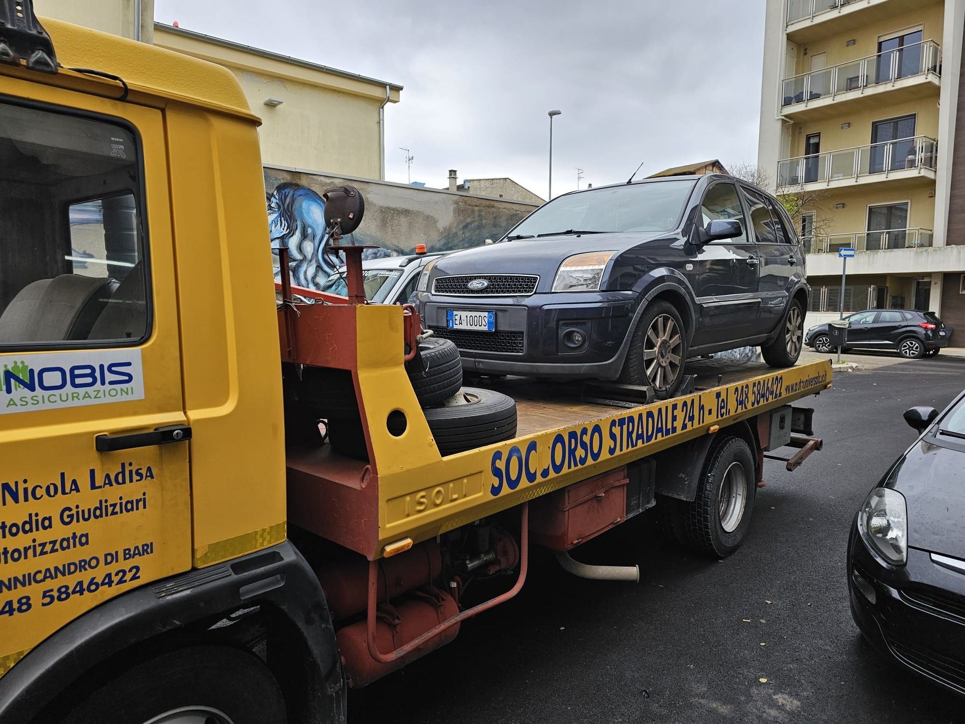 Un SUV Ford grigio scuro caricato su un carro attrezzi giallo con pianale, in una strada cittadina.