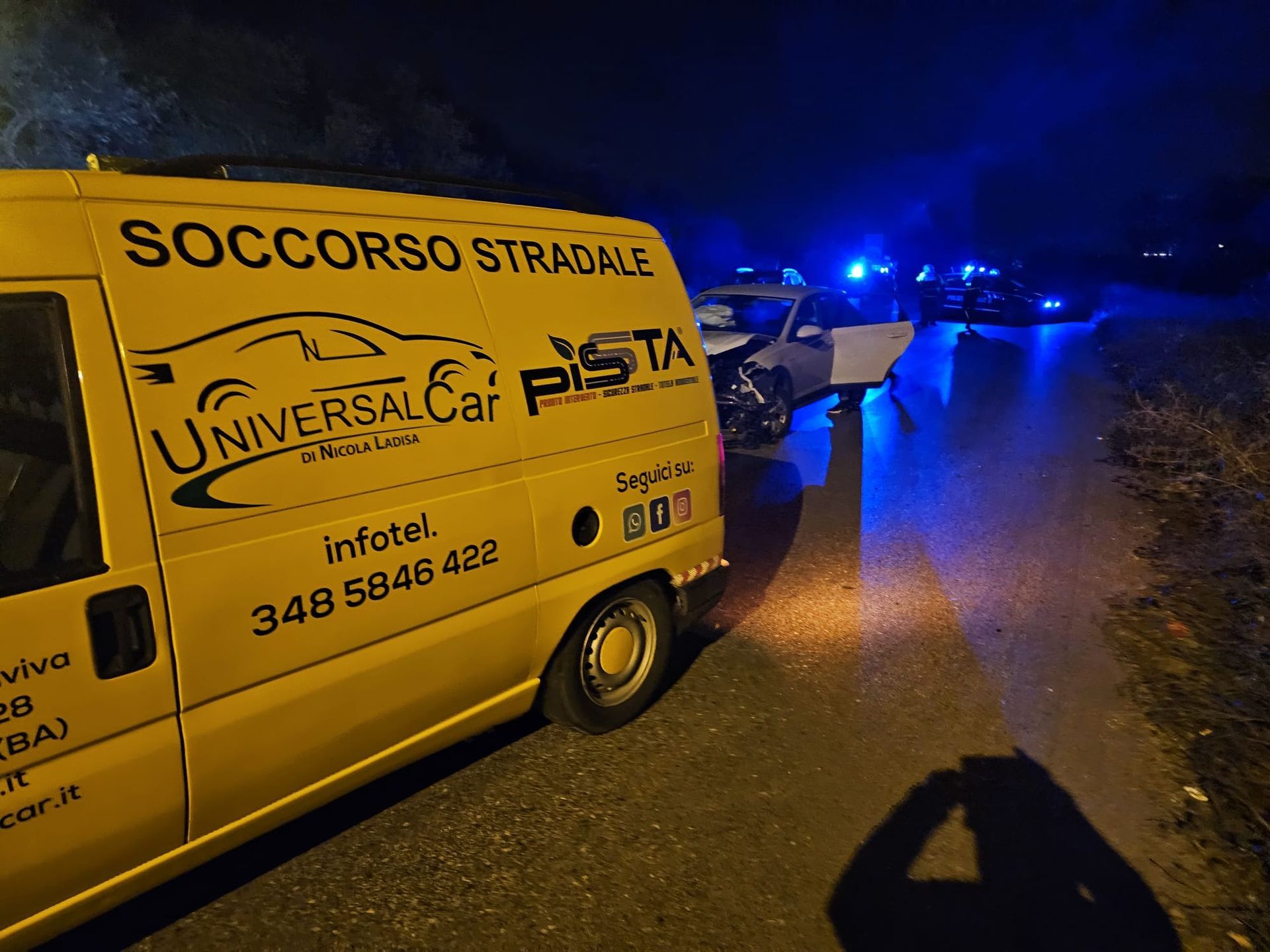 Un furgone giallo del soccorso stradale parcheggiato di notte vicino a un'auto con la parte anteriore danneggiata, illuminato da luci di emergenza blu.