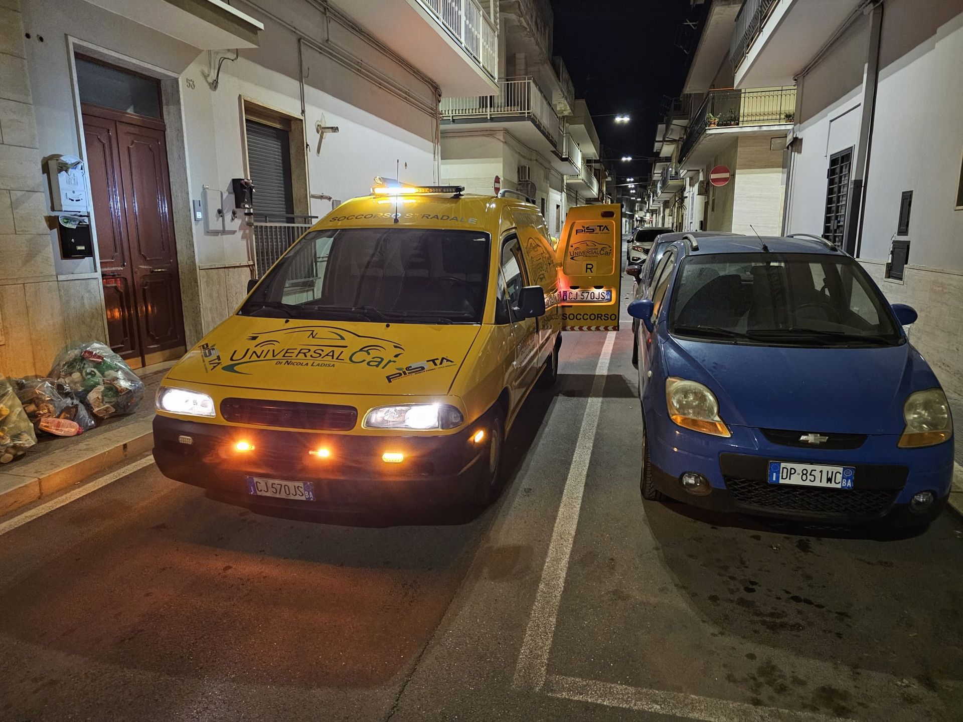 Un furgone giallo e un'auto blu parcheggiati di notte in una strada stretta, con una pila di borse sul marciapiede.