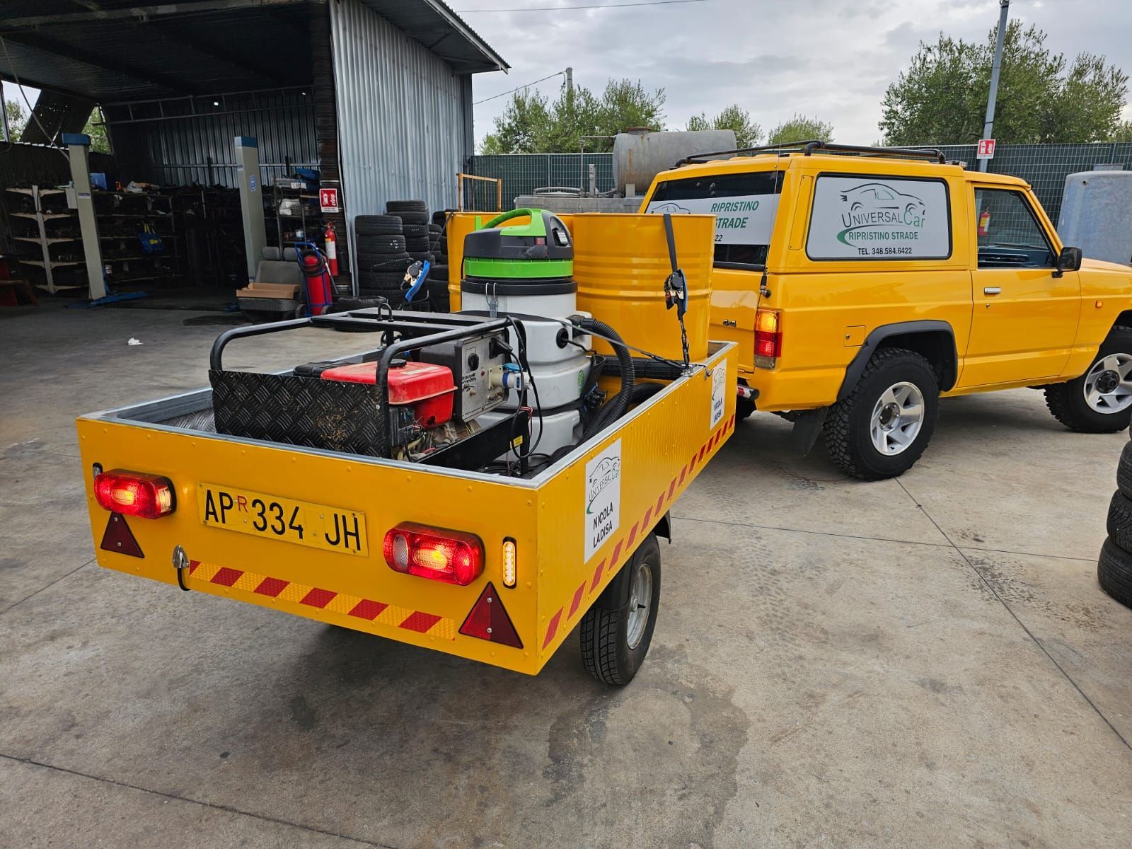 Un rimorchio giallo con attrezzature, agganciato a un SUV giallo, parcheggiato su un piazzale asfaltato presso un'officina.