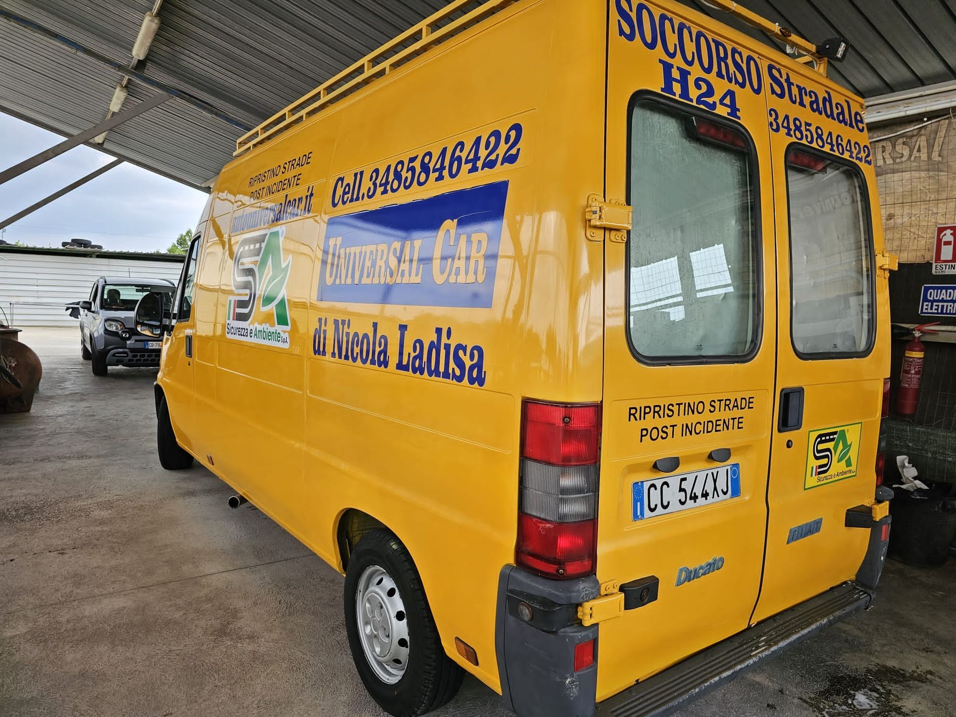 Un carro attrezzi giallo della Universal Car parcheggiato sotto una tettoia, con il nome dell'azienda, il numero di telefono e la targa ben visibili.