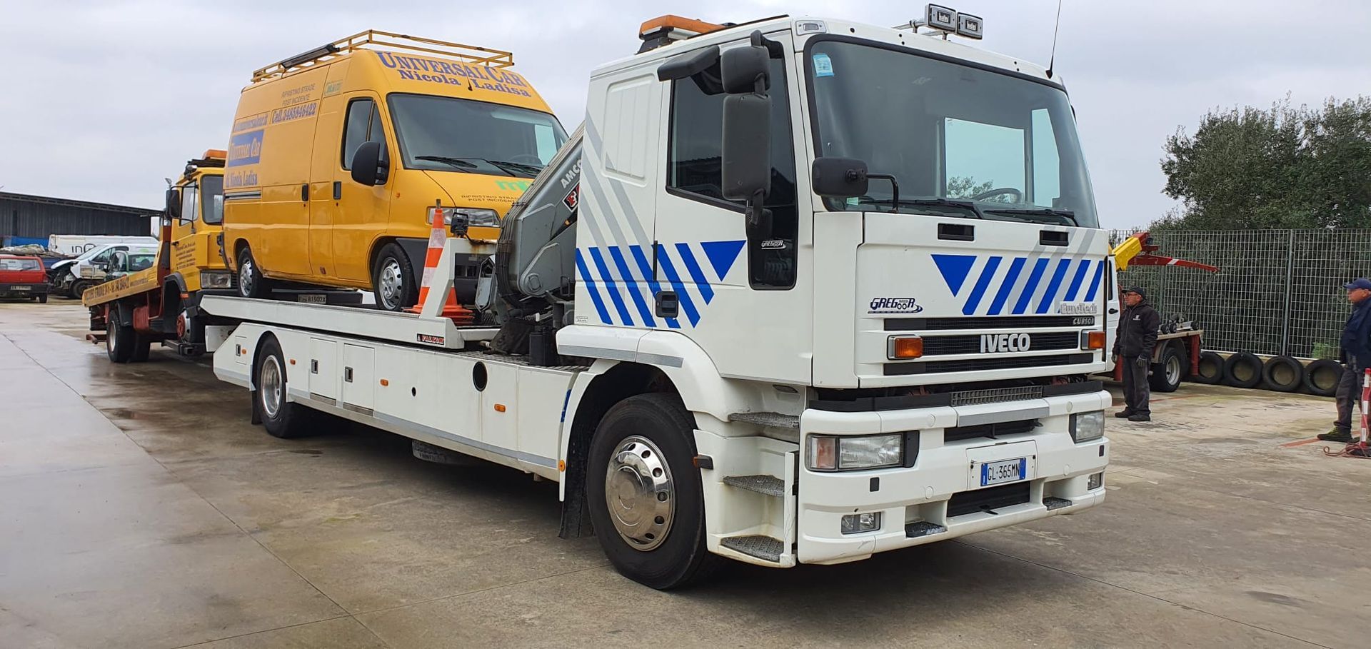 Un carro attrezzi bianco con pianale e decorazioni a chevron blu, che trasporta un furgone giallo, in un piazzale ghiaioso sotto un cielo nuvoloso.
