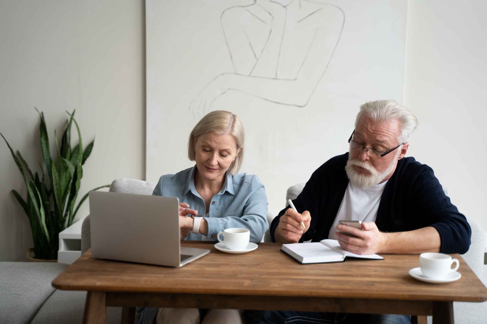 An elderly couple is sitting at a table with a laptop and a cell phone.