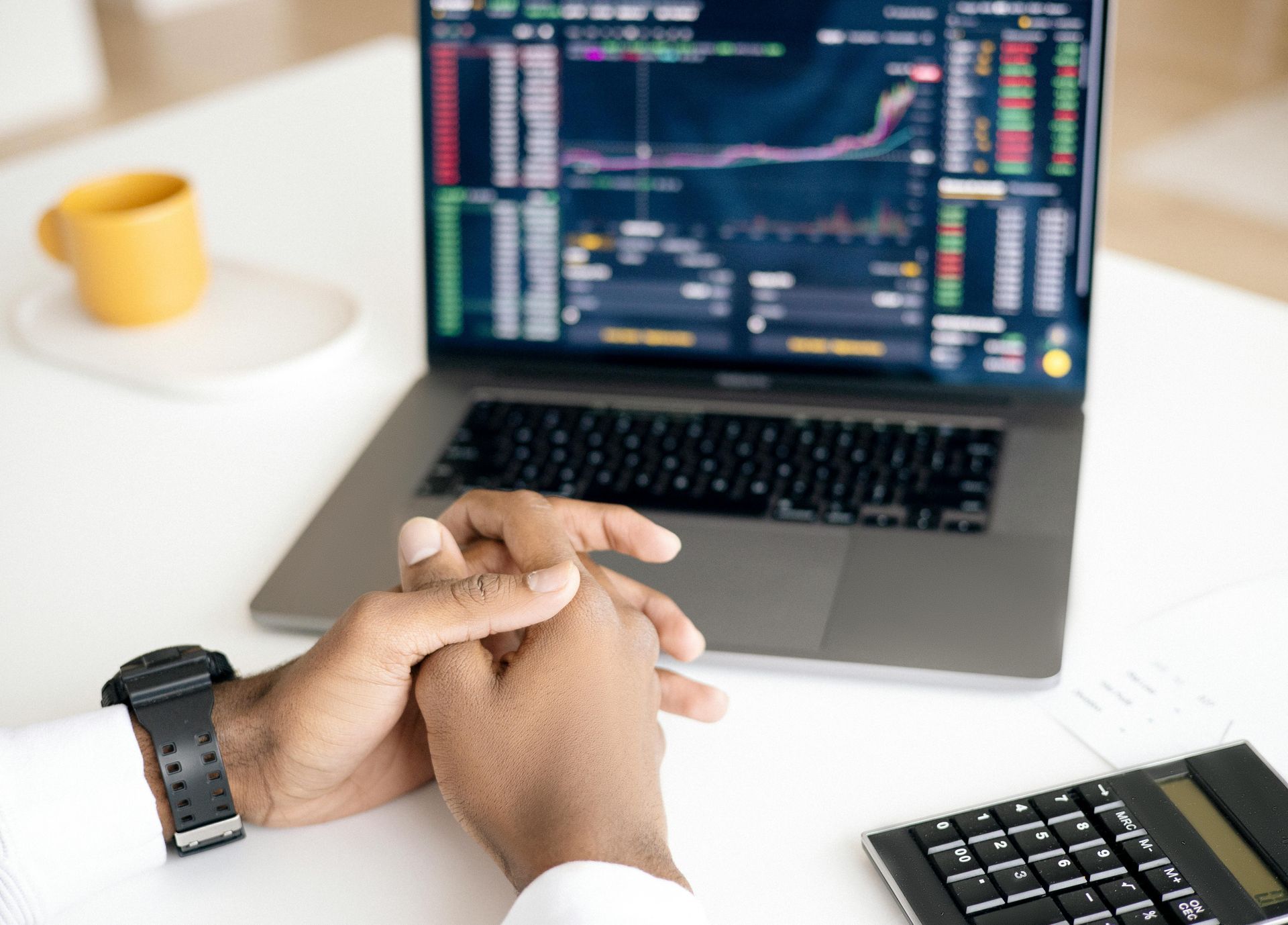 Person's hands clasped near a laptop displaying stock market charts, with a calculator and coffee cup.