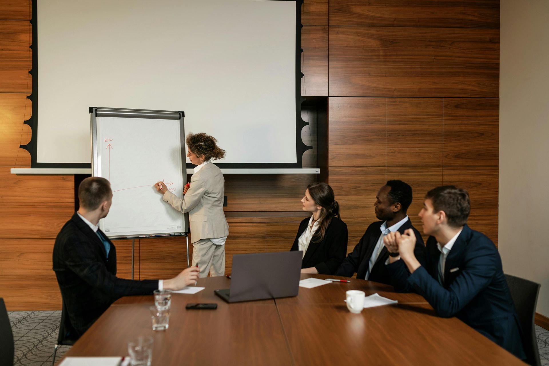 Business meeting: a person writes on a whiteboard while colleagues observe around a table in a wood-paneled room.