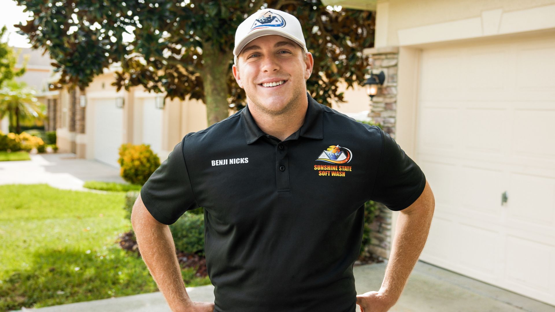 Man in black polo, hands on hips, smiling in front of a house, wearing a hat with a logo.