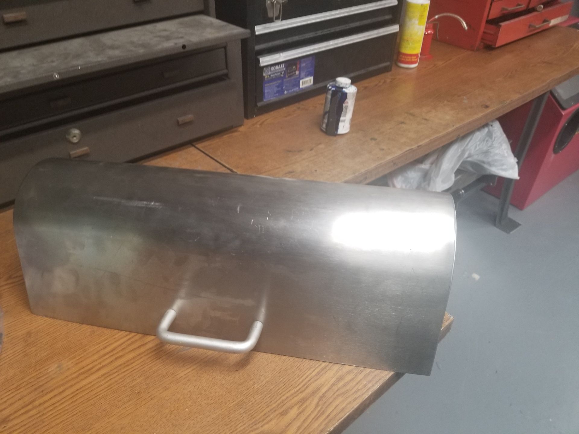 Stainless steel rectangular container with a handle sits on a wooden workbench in a workshop.