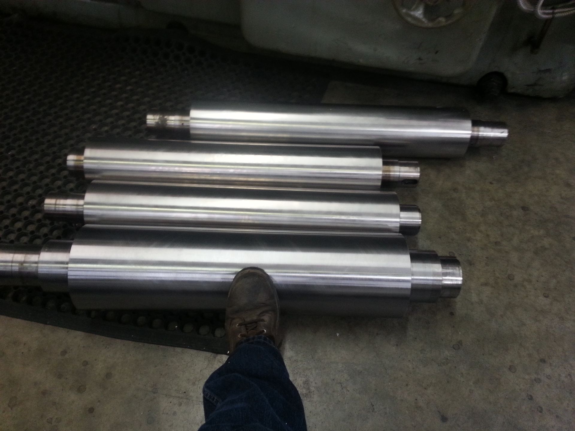 Four cylindrical metal rollers with shiny surfaces, near a person's foot, on a factory floor.