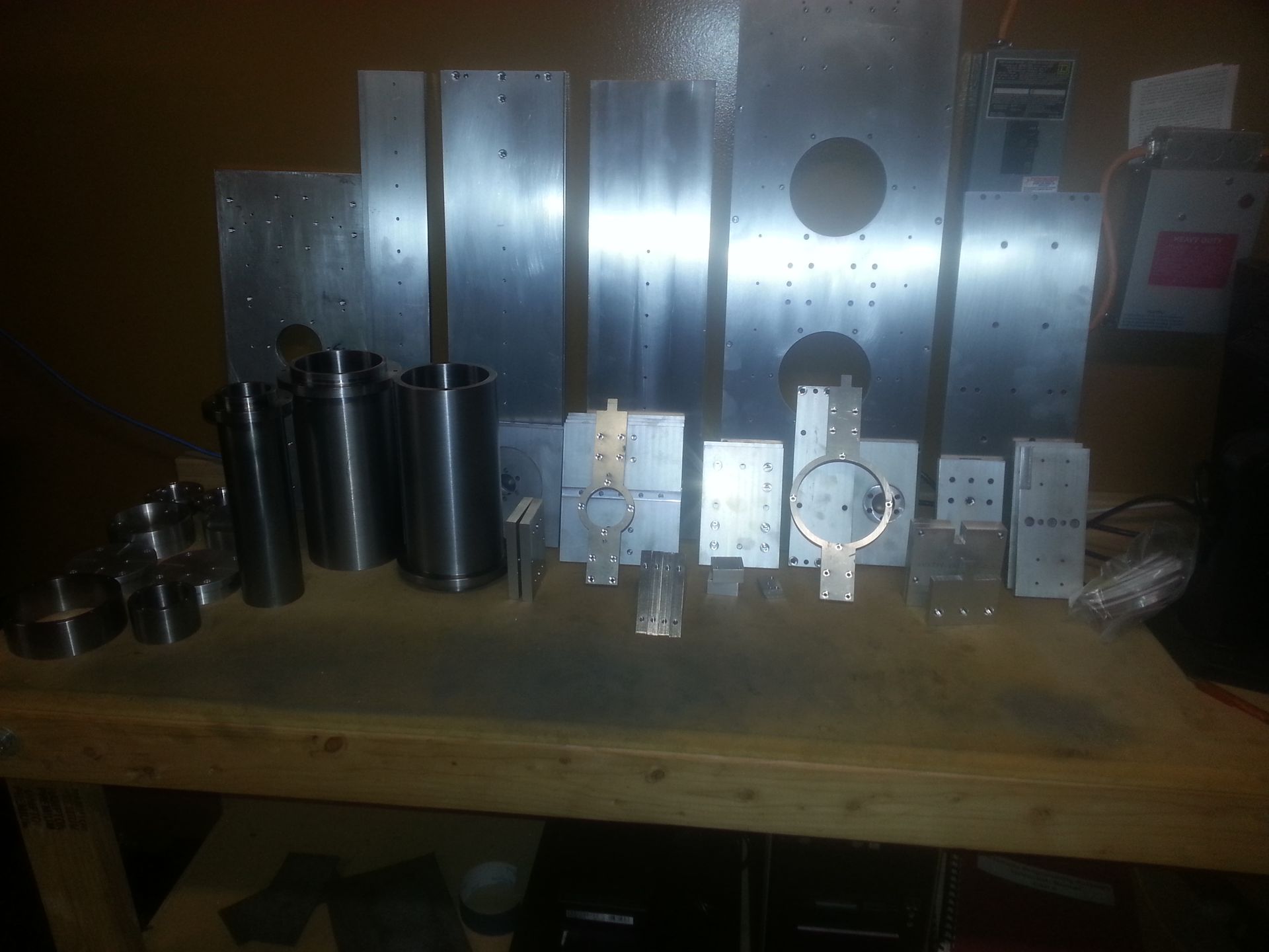 Metal parts arranged on a wooden surface. Cylindrical tubes and rectangular pieces with drilled holes are visible.