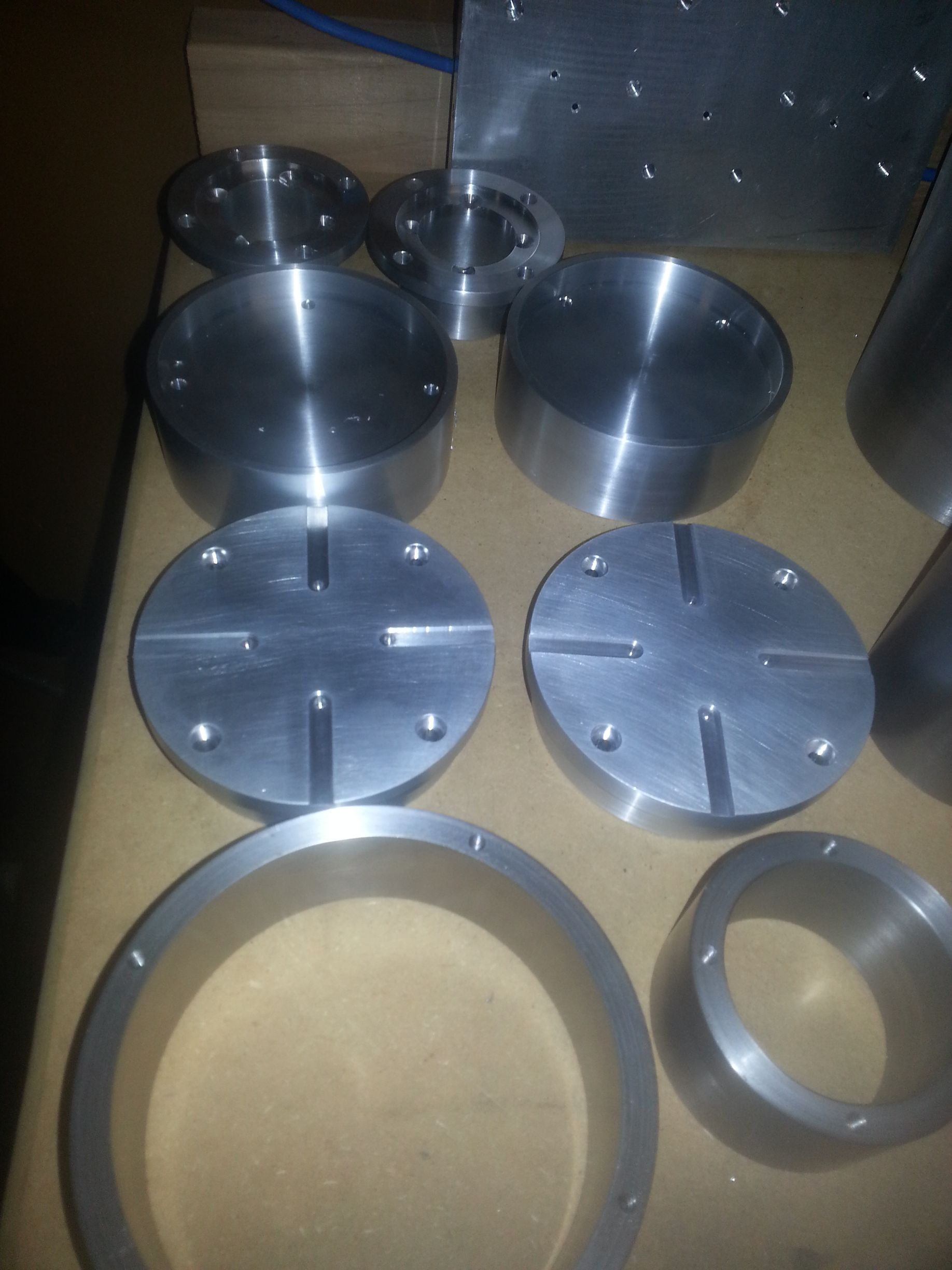 Machined metal parts, various shapes: cylinders, rings, plates, and a conical shape, arranged on a surface.