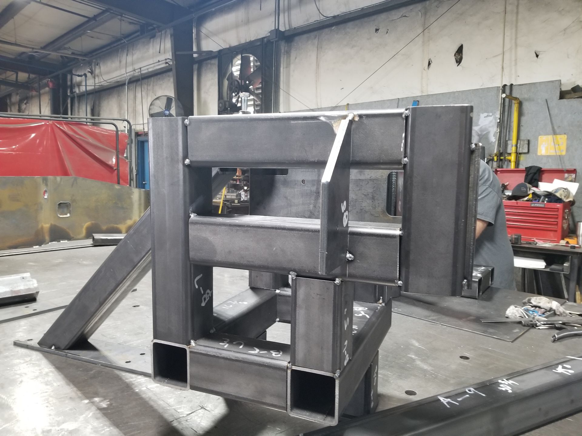 Welded steel frame in a workshop, under construction. A person is partially visible working nearby.