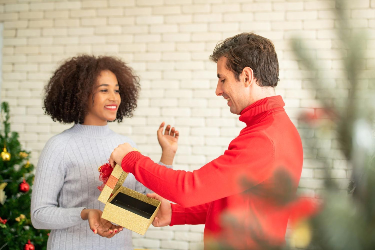 Em uma festa, uma pessoa vestindo um suéter cinza entrega uma caixa de presente dourada para outra de suéter vermelho.