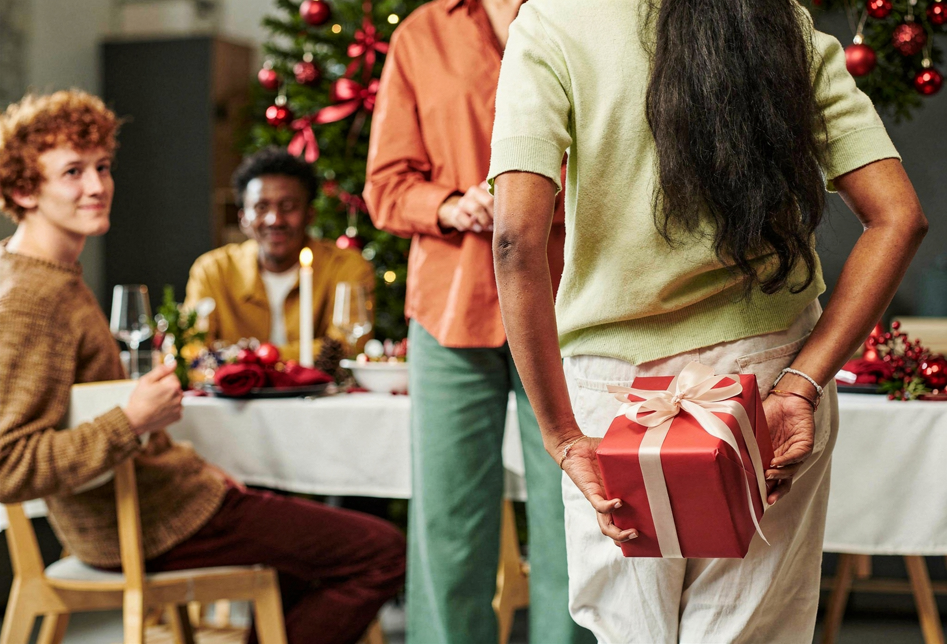 Uma pessoa segura uma caixa de presente vermelha atrás das costas, em pé diante de uma mesa de jantar com outras pessoas.