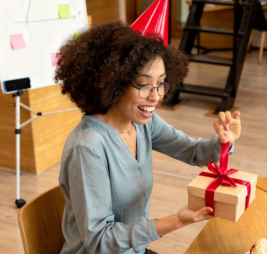 Uma pessoa com chapéu de festa sorri ao abrir uma pequena caixa de presentes com fita vermelha num ambiente de escritório