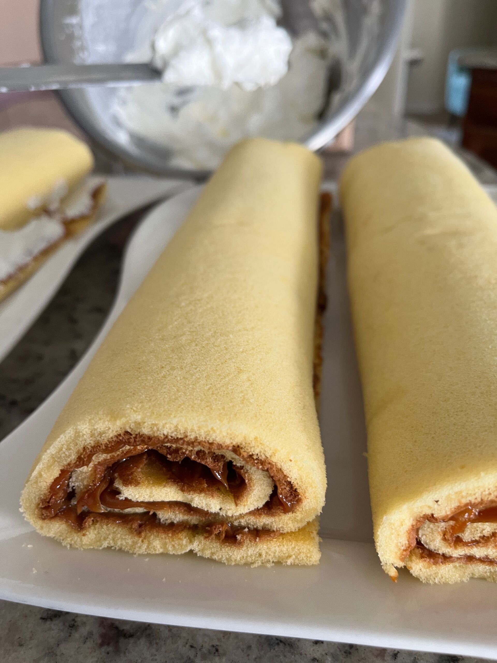 Two filled cake rolls on a white plate; creamy filling is being spread.