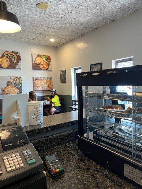 Interior of a cafe. Cash register and pastry display in foreground. Customers seated at a booth, food art on the wall.