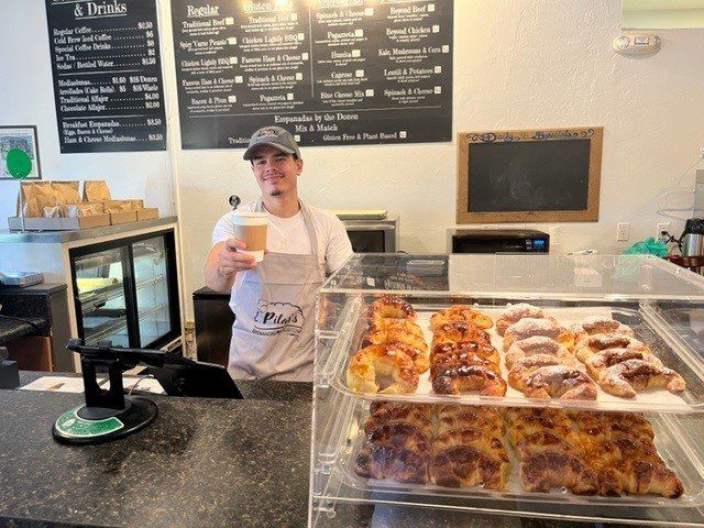 Man in apron holds coffee at a bakery counter with pastries, shop menu, and cash register.