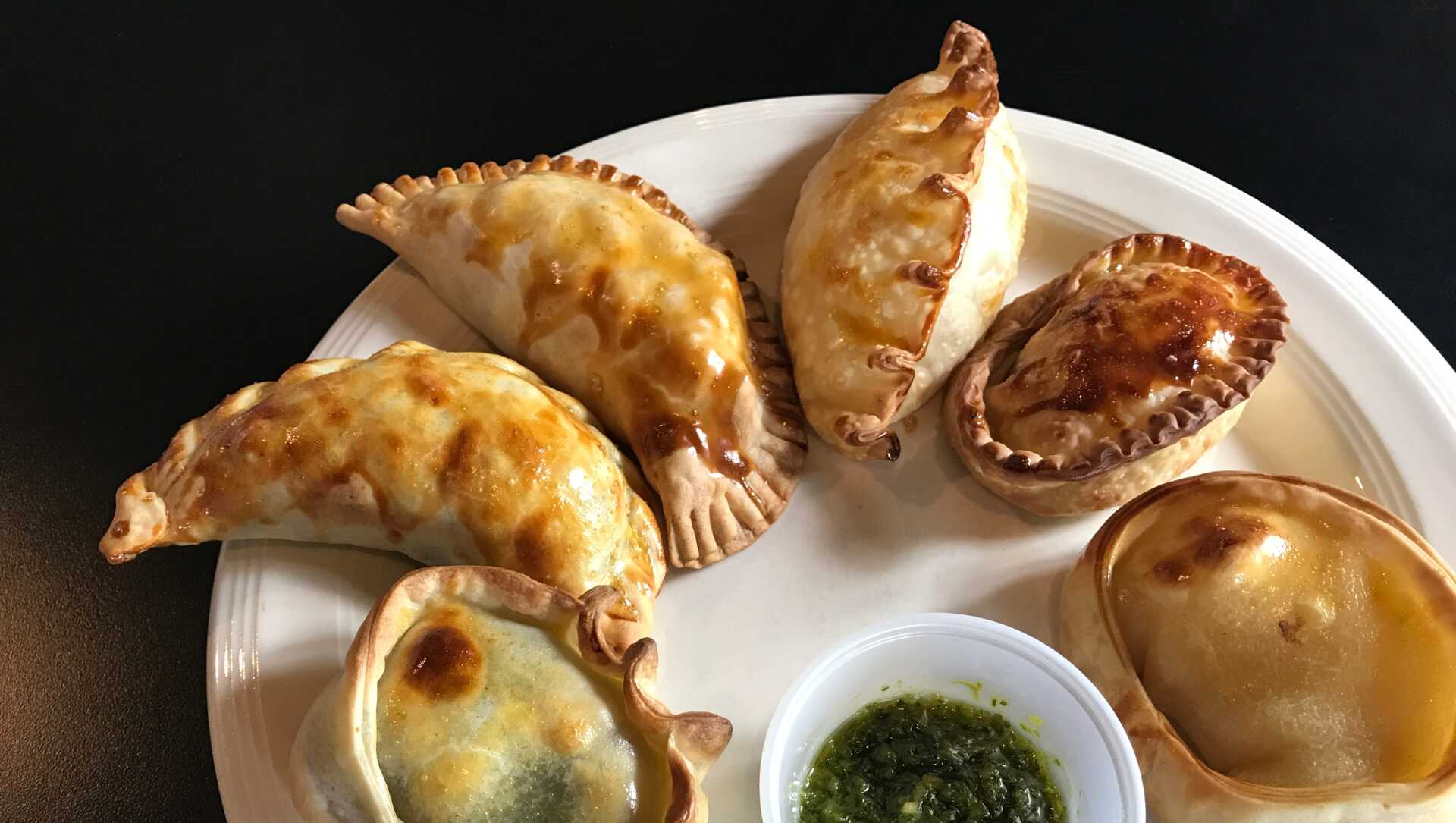 Plate of golden-brown empanadas with a small cup of green chimichurri sauce.